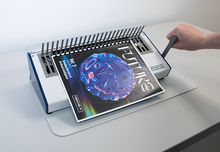 Load image into Gallery viewer, A white and blue binding machine on a light gray table, with a spiral-bound book or magazine being fed through the rollers. A human hand is visible holding the handle, and the book cover shows a blue reflective orb design with the word 'FUTURES'.