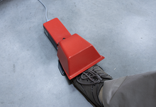 Load image into Gallery viewer, A red and black industrial foot pedal device with a gray shoe resting on its platform, set on a gray concrete floor.