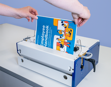 Load image into Gallery viewer, A person's hands are shown inserting a colorful 'Employee Handbook' booklet into a white and blue document binding machine on a light gray tabletop against a blue background.