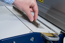 Load image into Gallery viewer, A close-up of a person's hand adjusting a row of metal guide bars on a white and blue binding machine.