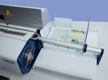 Load image into Gallery viewer, A close-up of a white and blue binding machine with a metal roller guide and a printed sheet being fed through. The machine has a visible warning label and a blue fan housing, photographed on a clean white surface against a pale blue background.