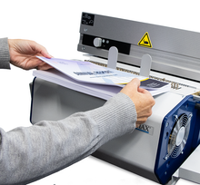 Load image into Gallery viewer, A close-up of a person's hands feeding a stack of printed documents into a  binding machine. The machine has a yellow warning label and a visible cooling fan on the side.