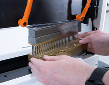Load image into Gallery viewer, A close-up of a person's hands positioning a brass template with a row of small pins into a metal press machine with orange handles.
